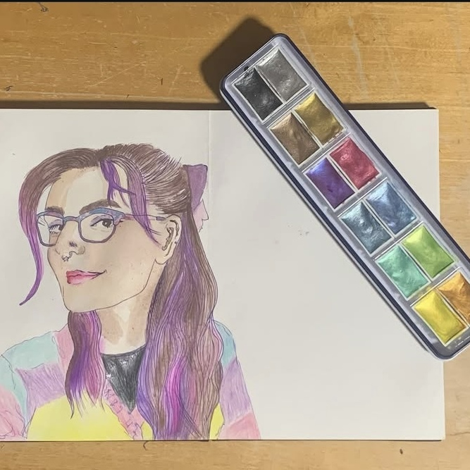 A watercolour painting on a whie, landscape page is on a brown desk with the glittery pallet used to paint some of it, open and horizontal across the top right hand corner. The casing is dark blue. The painting is lined in fineliner and is on the left hand side of the page. It is a portrait of the illustrator. She has long, purple and brown hair, with a bow peeking out of the back and the hair texture has been emphasised with fineliner lines. She is seen from the shoulders up and is wearing a colourful, pastel but bright cardigan with her top slighly peeking out with a round neck, near the centre of her neck. Her face is off to her left, our righut slighlty so it is at a 3/4 angle, but she is still looking at the viewer.
